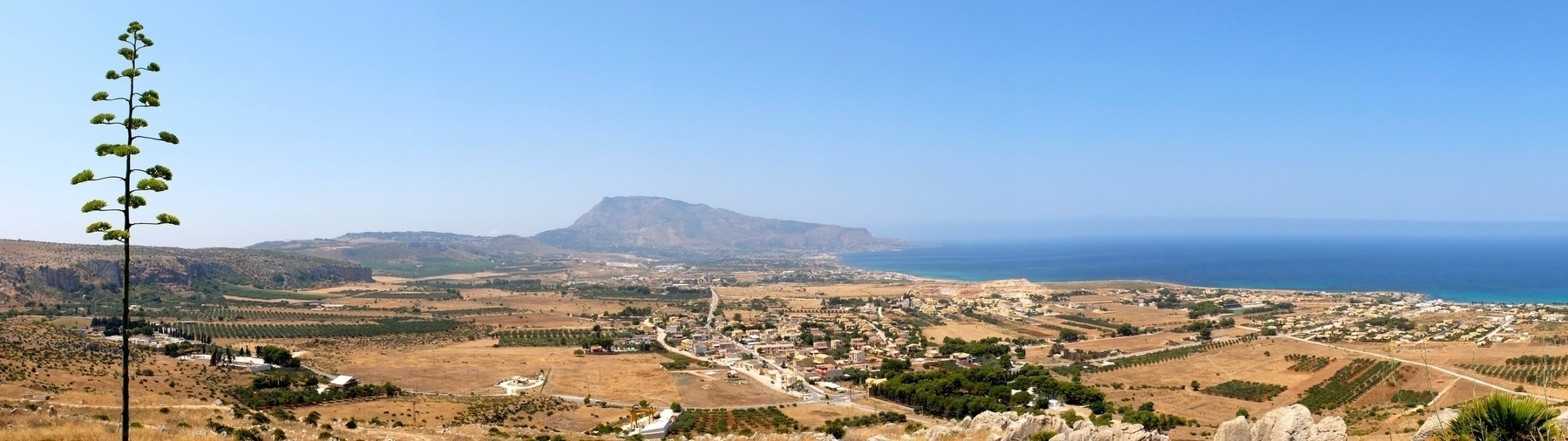 Une vue panoramique montre un paysage côtier avec des champs, des habitations, et une montagne en arrière-plan sous un ciel dégagé.