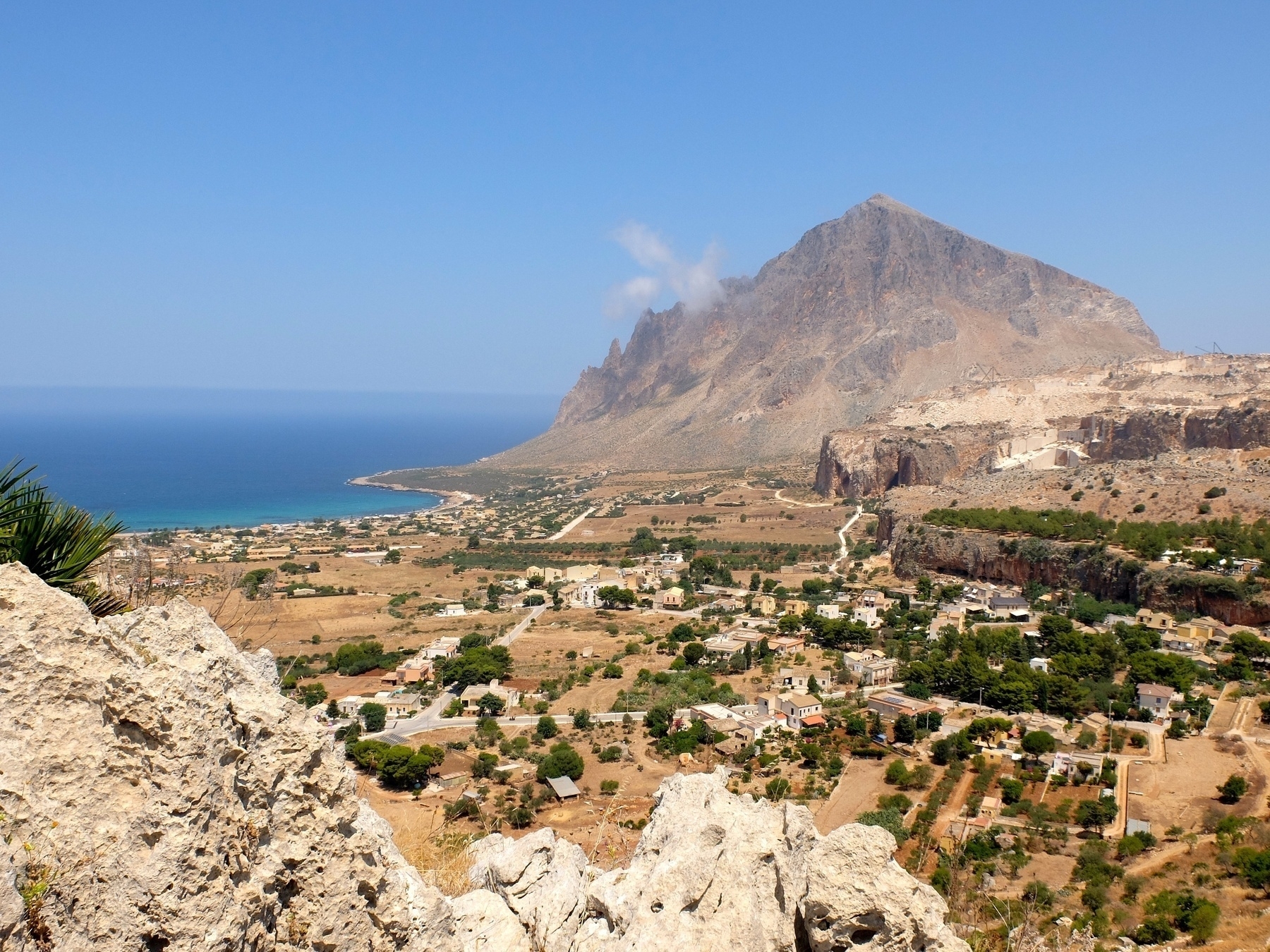 Une montagne majestueuse surplombe un paysage côtier méditerranéen avec des maisons et une végétation éparse.