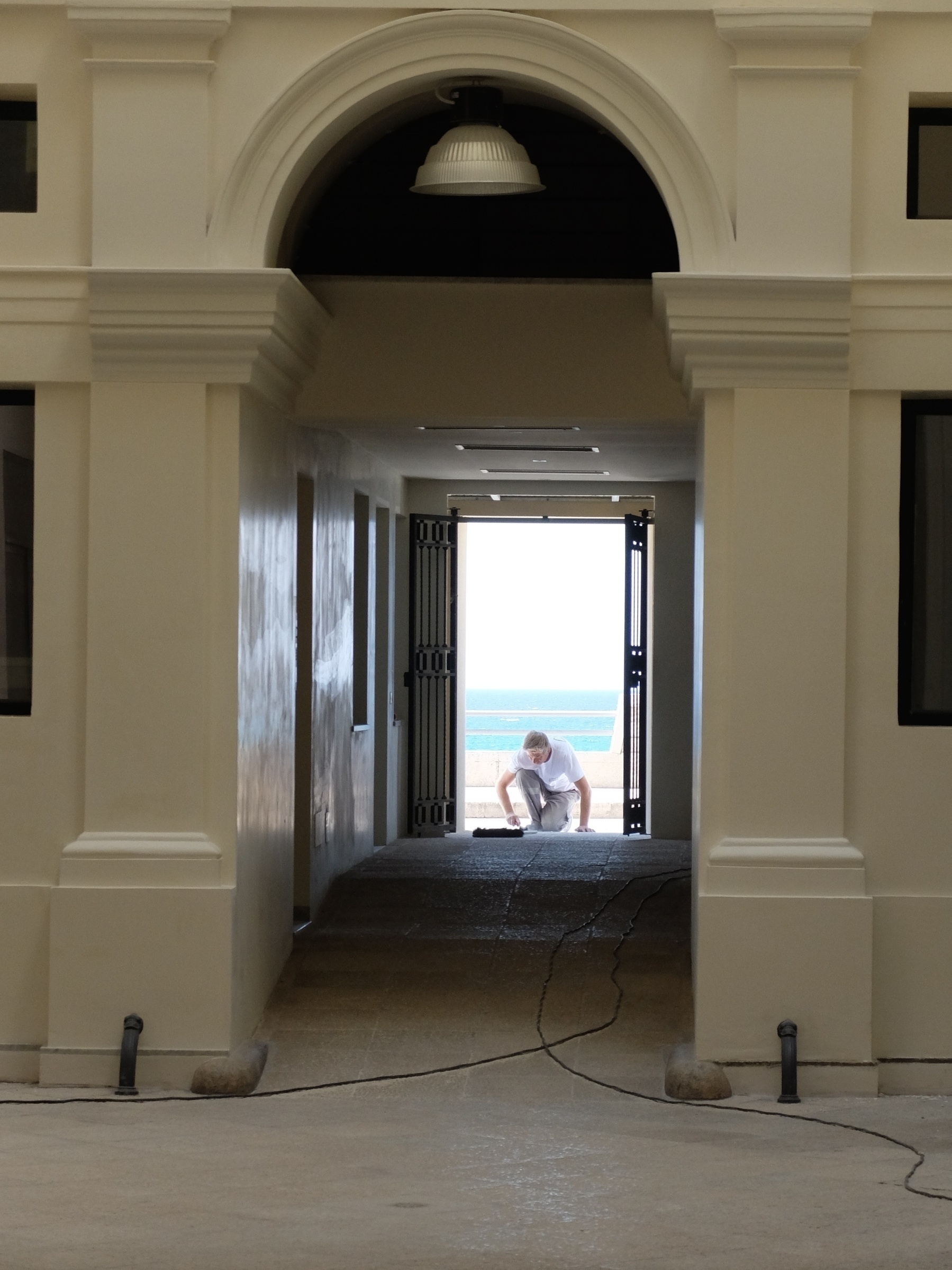 Un homme est accroupi à l'extrémité d'un couloir bordé d'arcades, avec une vue sur la mer en arrière-plan.