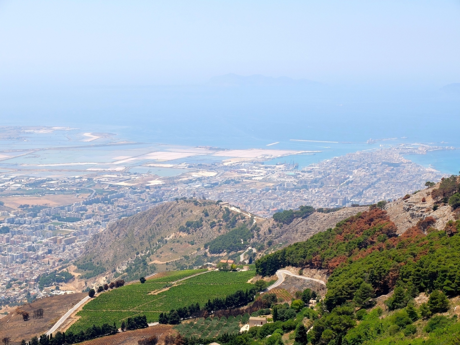 Une vue panoramique montre une ville côtière entourée de collines verdoyantes et de la mer en arrière-plan.